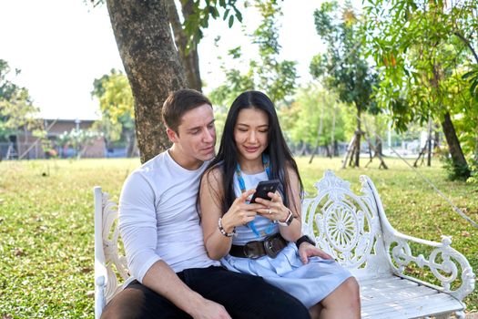 Newlywed multiracial couple sitting on a white bench in a park using a mobile phone during sunset
