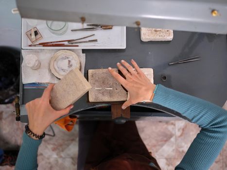 Top view of an unrecognizable woman working in her artisan jewelry workshop sitting at a workbench using stone blocks for her work.