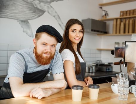 Coffee Business Concept - Positive young bearded man and beautiful attractive lady barista couple in apron looking at camera while standing at bar Couter ready to give Coffee Service at the modern coffee shop