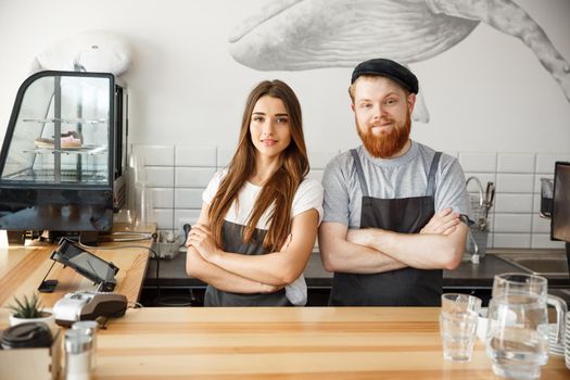 Coffee Business Concept - Positive young bearded man and beautiful attractive lady barista couple in apron looking at camera while standing at bar Couter ready to give Coffee Service at the modern coffee shop