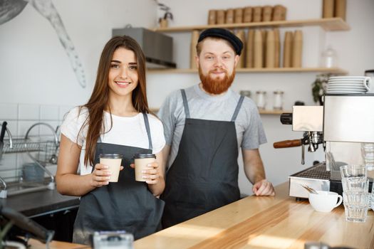 Coffee Business Concept - Positive young bearded man and beautiful attractive lady barista couple giving take away cup of coffee to custome at the modern coffee shop