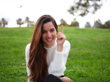 I have an idea. Front view of a young woman in the park looking at the camera smiling and raising her index finger. Concept of emotions.