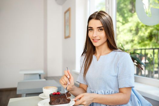 Young attractive Caucasian lady enjoy eating chocolate cake with hot coffee in modern coffee shop at noon.