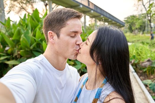 Portrait of a multiracial couple kissing while taking a selfie in a park during sunset
