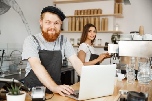 Coffee Business Concept - Positive young bearded man and beautiful attractive lady barista couple in apron looking at camera while standing at bar Couter ready to give Coffee Service at the modern coffee shop