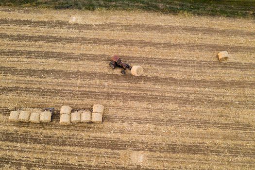 Aerial view from drone to the tractor collect bales of hay after harvesting on a wheat field.