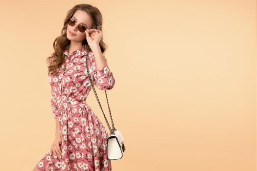 Studio portrait of stylish brunette young girl in sunglasses wearing nice summer dress with flower pattern and looking at camera. She is carrying a small white bag on chain and holding her skirt. Isolate.