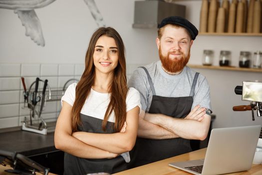 Coffee Business Concept - Positive young bearded man and beautiful attractive lady barista couple in apron looking at camera while standing at bar Couter ready to give Coffee Service at the modern coffee shop