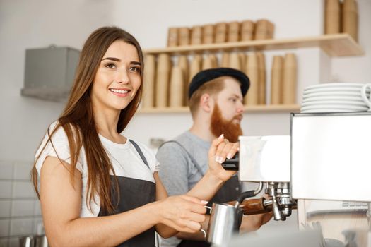 Coffee Business Concept - portrait of lady barista in apron preparing and steaming milk for coffee order with her partner while standing at cafe.