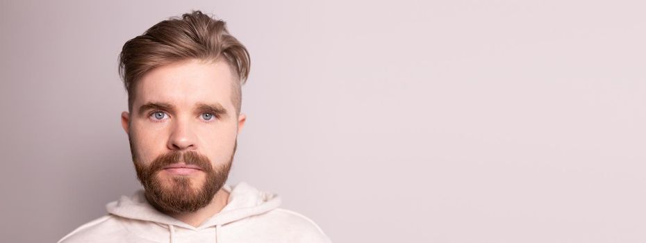 Portrait of young male with beard and mustache and trendy hairdo. Wears casual hoodie, has serious expression in studio against white