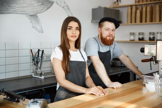 Coffee Business Concept - Positive young bearded man and beautiful attractive lady barista couple in apron looking at camera while standing at bar Couter ready to give Coffee Service at the modern coffee shop