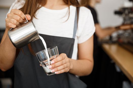 Coffee Business Concept - close-up lady barista in apron preparing and pouring milk in glass cup while standing at cafe.