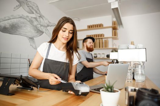 Coffee Business Concept - Beautiful female barista giving payment service for customer with credit card and smiling while working at the bar counter in modern coffee shop.