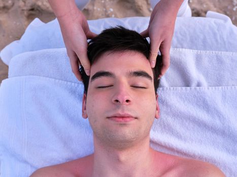 close up view from above of a young man lying on a massage table receiving a head massage on a beach in Valencia by a young chiromassage therapist.