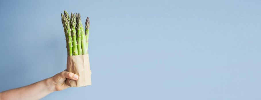 Female hand holds Bundle of green asparagus on blue background. Concept of vegans, vegetarians and healthy food.