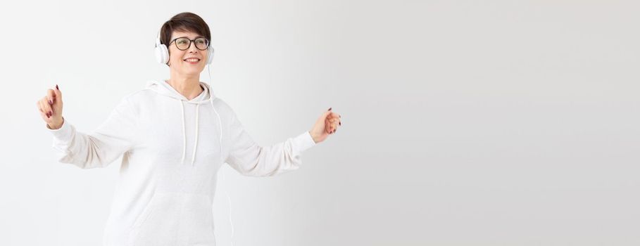 Pretty keen middle-aged woman in glasses and a white sweater listens to her favorite music with headphones on a white background. Online radio and music subscription concept. Copyspace