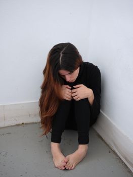 Vertical image of a young woman in the corner of a room dressed in black barefoot looking down in desolation. Concept of emotions.