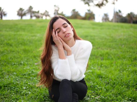 Front view of a young woman in the park sitting on the grass with her hands on her face and her eyes closed daydreaming. Concept of emotions.