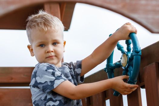 Beautiful child is driving a ship wheel in a playground. Kid wearing camouflage feels relaxed in a wooden playhouse. Childhood full of adventure