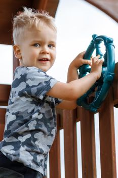 Cute child is driving a ship wheel in a playground. Kid wearing camouflage feels wild in a wooden playhouse. Childhood full of adventure. Wind tangled hair