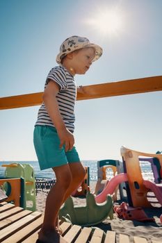 A cute boy in a hat walks on a wooden bridge to the beach. A playground, sea and sun rays in the background