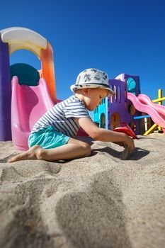 A little boy digs sand with his hands on the beach, a colourful playground visible in the background