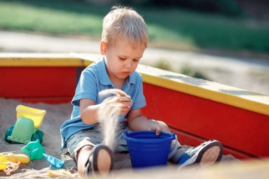 Toddler boy is playing in a red and yellow edge sandbox with sand toys. Focused baby, wearing blue shirt is trying to put sand in a blue bucket. Childhood games outdoor