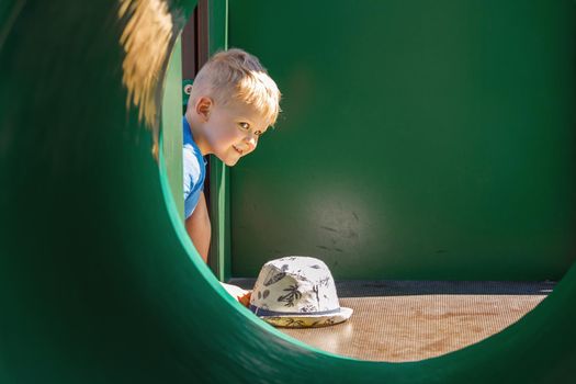 Child playing on Outdoor children green playground in a city park. The little boy takes off his hat, and looks at the camera through the green tunnel.