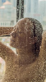 Beautiful woman in the shower behind glass with drops on the background of a window with a panoramic view of the city. VERTICAL FORMAT for Instagram mobile story or stories size. Mobile wallpaper