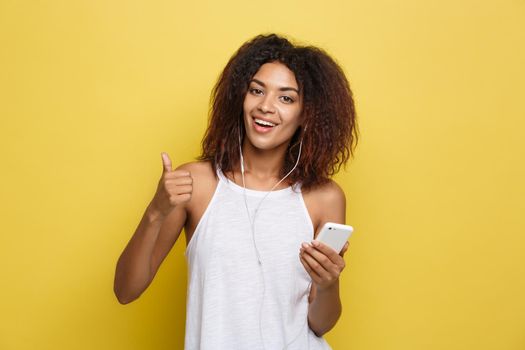 Lifestyle Concept - Portrait of beautiful African American woman joyful listening to music on mobile phone and show ok sign with fingers. Yellow pastel studio background. Copy Space