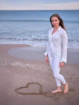 Vertical image of a young woman in white standing on the beach with a heart drawn in the sand looking at the camera.