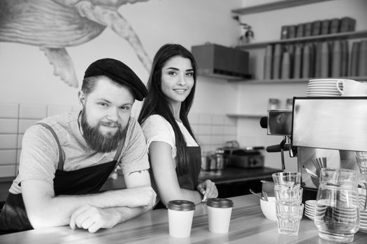 Coffee Business Concept - Positive young bearded man and beautiful attractive lady barista couple in apron looking at camera while standing at bar Counter.