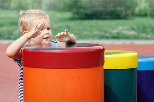 Small boy  plays a colorful drums in public playground. Happy child hiding behind a line of drums. Feel the music everywhere