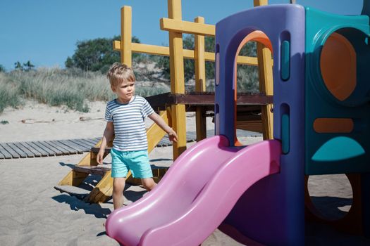 A little boy in a striped shirt on a beach playground near the pink slide