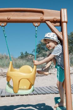 The cute little boy tries to climb into the yellow plastic swing himself. Independent, naughty child. Beach background
