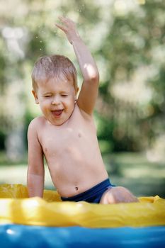 Very happy kid splashing water on himself head. Cold water and hot summer day. Enjoying water in the garden. Opened mouth and raised hand. Green nature background