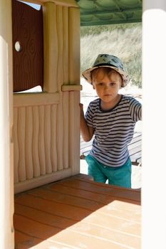 A boy in a hat and striped shirt poses on a beach playground looking straight at the camera