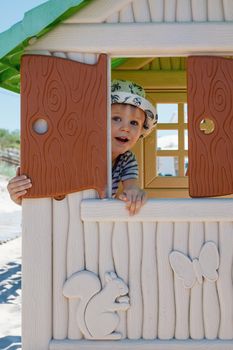 A cute boy is hiding in a small toy house, he is watching through an open shutter