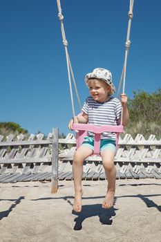 A cute boy swings on the beach. Pink plastic swing, blue sky and dune background