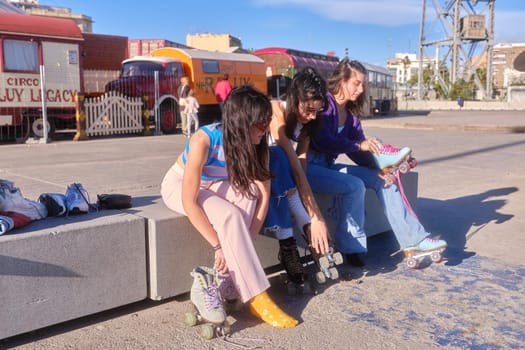 Three young women in retro style clothes sitting in a city park tying on classic skates