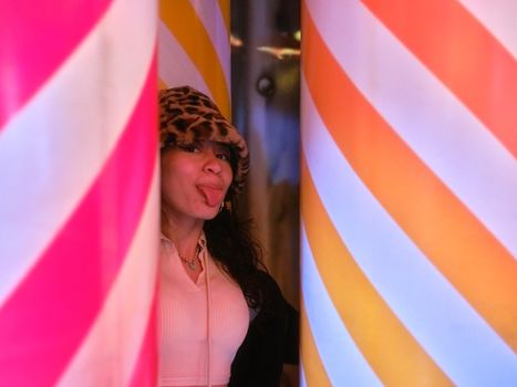 Latina woman in leopard hat sticking out her tongue joking between coloured columns at a night fair