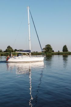 A white yacht with lowered sails and Lithuania flag in the lagoon. Quiet evening, well visible reflection of the ship's mast in the water. Vertical photo