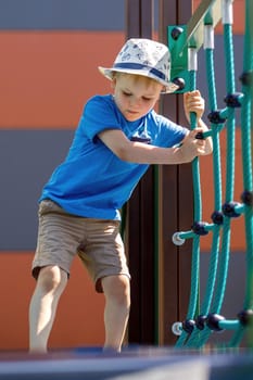 Low angle view of adorable little boy with hat having fun at playground. The child trains and carefully crosses the monkey bridge.