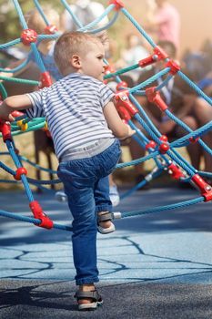 Cute boy having fun on outdoor playground. Summer active leisure for kids. Activity for family with children. Equipment of entertainment park for kids. The child is ready to climb the rope tower.