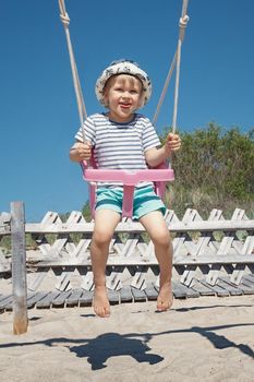 The little boy sways in the beach swing. The child is very cheerful, and naughty he shows his tongue