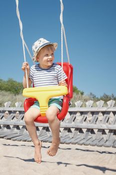 The beach, the blue sky and the happy boy swings in a red-yellow swing. The child smiles and looks to the side, there is free space for text