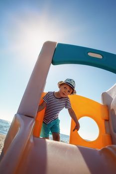 A little boy in a hat plays on a slider he climbs up. Sky, sun and sea in the background