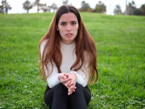 Front view of an angry young woman in the park sitting on the grass with her hands together looking at the camera. Concept of emotions.