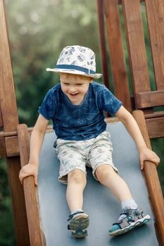 Cute baby boy riding on metallic slide at childish playground relaxing having summer vacation