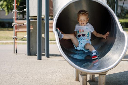 Happy Kid Sliding Fast In Tube Slide On Playground Park. Joyful Boy Having Fun in Play Centre. Kindergarten For Elementary Age Children.